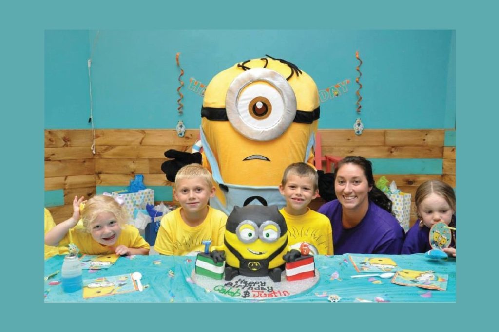 Showing a picture of mom and kids at a minion birthday party at Cowabunga's.  All are happy around a nicely decorated table with a minion character in the back.